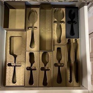 May include: A drawer filled with gold-colored kitchen utensils, including spoons, spatulas, and knives, each nestled in individual cardboard packaging. The utensils are arranged neatly, suggesting a set. The drawer is made of wood.