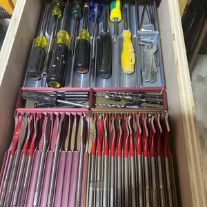 May include: A wooden drawer filled with various hand tools. The top section holds screwdrivers with black and yellow handles. Below are sets of drill bits and red-handled spade bits, neatly organized in pink and gold cases. A practical assortment for any workshop.