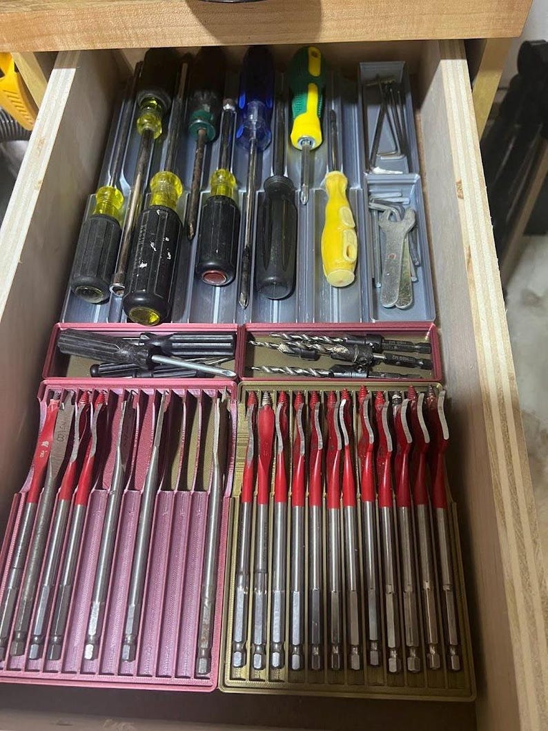 May include: A wooden drawer filled with various hand tools. The top section holds screwdrivers with black and yellow handles. Below are sets of drill bits and red-handled spade bits, neatly organized in pink and gold cases. A practical assortment for any workshop.