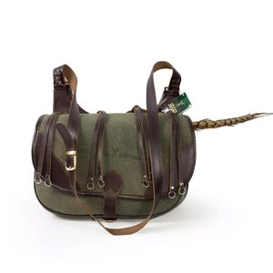 May include: A green canvas and brown leather hunting bag with a pheasant feather attached to the side. The bag has multiple straps and buckles.