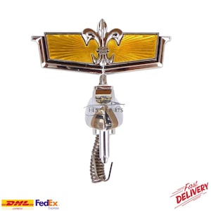 May include: A chrome and gold car emblem featuring a fleur-de-lis design. The emblem has a rectangular shape with a sunburst pattern. The text "HLS CAR PARTS" is visible on the emblem.