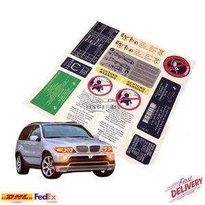 Op de afbeelding: Een zilveren BMW X5 SUV met een set waarschuwingsstickers. De stickers hebben tekst en afbeeldingen, waaronder "ACHTUNG WARNING" en airbagwaarschuwingen. De afbeelding bevat ook de logo's van DHL en FedEx.