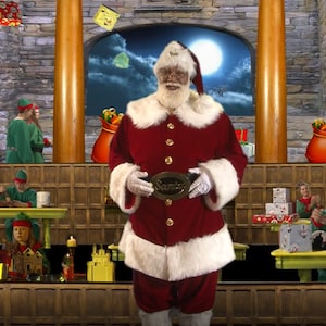 Puede incluir: Santa Claus, vestido con un traje rojo con ribetes blancos, sostiene una hebilla de cinturón dorada que dice "Santa". Está de pie frente a una pared de ladrillo con un árbol de Navidad a cada lado. Hay duendes trabajando en el fondo.