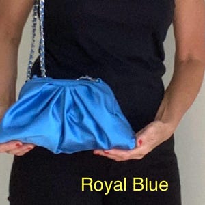 May include: A royal blue clutch bag with a gathered, pleated fabric design and a silver-toned chain strap. The bag features a soft, satin-like texture and a structured frame closure. The text Royal Blue is visible at the bottom of the image.