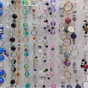 May include: A selection of beaded keychains in diverse colours, such as blue, black, clear, and pink. Each keychain displays a different number and decorative beads, with silver-toned metal rings and chains.