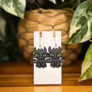 May include: A pair of dangle earrings featuring black dragon charms with large green eyes, small wings, and silver-toned metal hooks. The charms are shaped like cartoon creatures and are displayed on a white vertical stand for jewelry presentation.