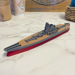 May include: A model battleship with a red hull, grey superstructure, and a wooden deck. The model has detailed features including turrets, guns, and other ship components. The ship is a toy or collectible item.