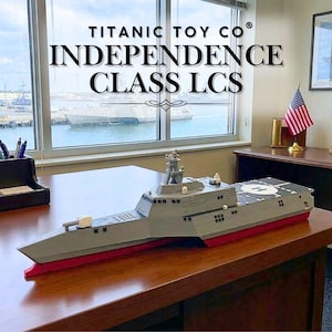 Puede incluir: Maqueta gris y roja de un buque de guerra LCS de clase Independence sobre un escritorio de madera. El modelo tiene detalles, incluyendo una plataforma para helicópteros y armamento. El texto "TITANIC TOY CO® INDEPENDENCE CLASS LCS" está visible.