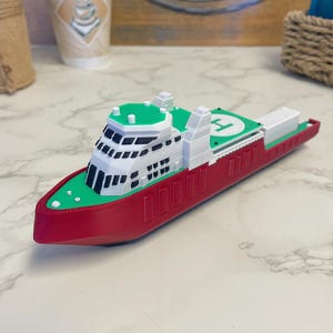 May include: A red, white, and green toy ship with a helipad on top. The toy ship features detailed design elements, including windows and a small white structure at the stern. The toy is likely made of plastic and resembles a real-life vessel.