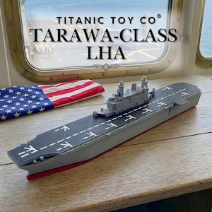May include: A gray and red model of a Tarawa-class LHA aircraft carrier, with white markings on the deck. An American flag is in the background. The text "TITANIC TOY CO" and "TARAWA-CLASS LHA" is visible.
