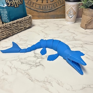 Puede incluir: Un juguete de dinosaurio articulado azul brillante, posiblemente un Mosasaurio, descansa sobre una superficie de mármol blanco. El juguete tiene un cuerpo segmentado, aletas y una cabeza detallada con la boca abierta. Un juguete divertido y educativo.
