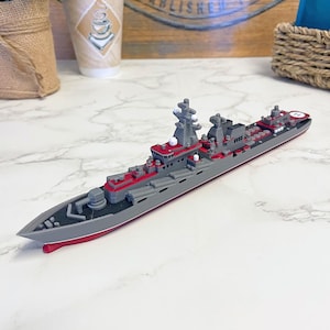 May include: A gray and red model warship with detailed features, including turrets, radar, and a helipad. The model has a streamlined design with a red waterline and is set against a neutral background.