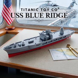以下が含まれることがあります： テーブルの上に置かれた、USS Blue Ridgeのグレーと赤色の玩具モデル。モデルには詳細な構造と赤い船体があります。背景にはアメリカ国旗と「Titanic Toy Co」と「USS BLUE RIDGE」の文字が表示されています。