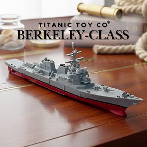 May include: A grey and red model warship, labelled "BERKELEY-CLASS" by "TITANIC TOY CO". The toy ship is detailed with various structures and weaponry, resting on a wooden surface. A nautical theme is suggested by the background.
