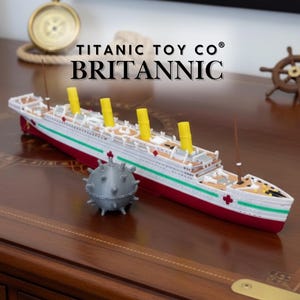 May include: A model of the Britannic ship with red, white, and green accents, and yellow smokestacks. The model is accompanied by a gray, spiky sphere. The text "TITANIC TOY CO® BRITANNIC" is displayed above the ship.