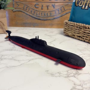 May include: A black and red submarine model featuring a conning tower with periscopes and a rear propeller. The hull is primarily matte black with a distinct red lower section, showcasing a detailed cylindrical shape with visible hatch markings.