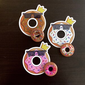 May include: Three donut stickers and three donut-shaped pins. The stickers feature a donut with sunglasses and a crown. The donuts have brown, white, and pink frosting with colorful sprinkles. The pins have similar designs.