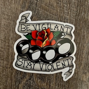 May include: A white sticker with a black and white illustration of a knuckle duster with a red rose on top. The sticker has the text "Be Vigilant Stay Violent" in a banner.
