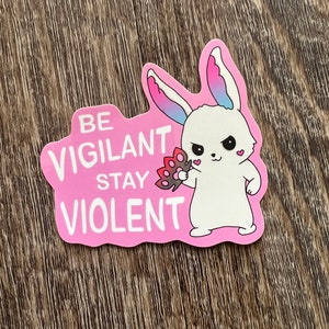 Puede incluir: Una pegatina rosa con un conejo blanco de dibujos animados que sostiene una flor. La pegatina dice "BE VIGILANT STAY VIOLENT". El conejo tiene detalles rosas y azules.