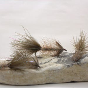 May include: Three brown and tan fishing flies with fine, wispy hackle fibres and thin, elongated bodies. The flies are arranged on a smooth, light-coloured, mottled stone with grey and beige patches, showcasing detailed feather textures and hooks.