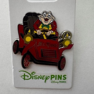 May include: A Disney Parks pin featuring Mr. Toad driving a red car. The pin has a black seat, yellow headlights, and the words "Mr. Toad" on the car. The pin is on a white backing card with the Disney Pins logo.