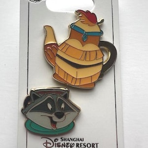 Puede incluir: Dos pines de esmalte de Disney en una tarjeta blanca. Un pin es una tetera con un tocado nativo americano. El otro pin es un mapache con un cuello verde. La tarjeta dice "Shanghai Disney Resort" en inglés y chino.
