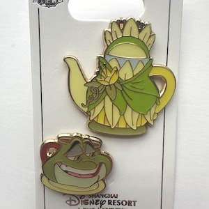 Puede incluir: Dos pines de esmalte de Disney con personajes de Tiana y el Sapo. Un pin es una tetera amarilla y verde, y el otro es una rana sonriente. Los pines están en una tarjeta blanca con el logotipo de Shanghai Disney Resort.