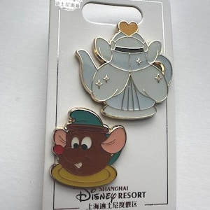 Puede incluir: Dos pines de esmalte de Disney en una tarjeta gris. Un pin es una tetera azul claro con ribetes dorados y un corazón en la parte superior. El otro pin es un ratón marrón en una taza. La tarjeta dice "Shanghai Disney Resort".