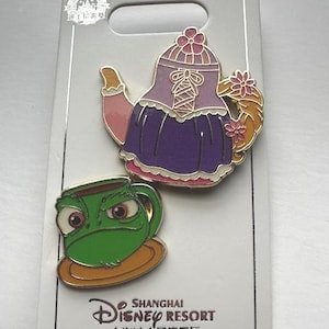 May include: Two enamel pins featuring Disney characters. One pin is a purple teapot with pink and gold accents. The other is a green frog-faced teacup on a gold saucer. The pins are on a white card with "Shanghai Disney Resort" text.