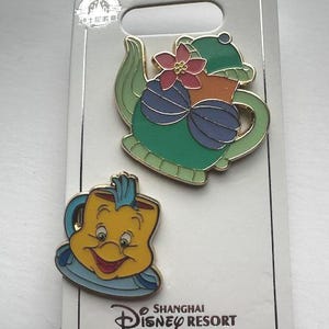May include: Two Disney enamel pins featuring characters from Beauty and the Beast. One pin is a green teapot with a pink flower, and the other is a yellow teacup with a blue rim. The pins are on a white card with the Shanghai Disney Resort logo.