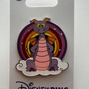 May include: A Disney pin featuring Figment the dragon, with a purple body, pink belly, and orange wings, set against a rainbow and cloud background. The pin is on a white card with the Disney Pins logo.