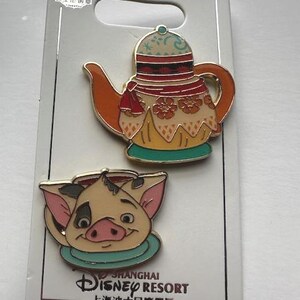 Könnte beinhalten: Zwei Disney-Emaille-Anstecknadeln auf einer weißen Karte. Eine Anstecknadel ist eine Teekanne mit orangefarbenen, roten und gelben Details. Die andere ist ein Schweinegesicht mit rosa Ohren und einem türkisfarbenen Kragen. Auf der Karte steht "Shanghai Disney Resort".