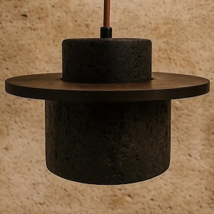 Puede incluir: Una moderna lámpara colgante con una pantalla cilíndrica oscura y texturizada y un acento circular de madera. La luz cuelga de un cable marrón. La lámpara tiene un diseño minimalista, adecuado para varios estilos de interior.