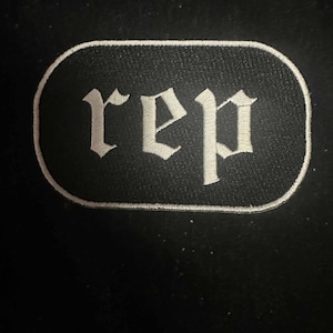 Può includere: Toppa ricamata bianca con la parola "rep" in carattere gotico su sfondo nero.