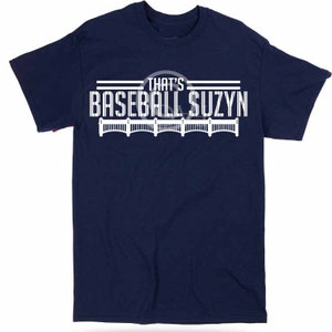 May include: A navy blue t-shirt with white text that reads "That's Baseball Suzyn" with a baseball graphic and a white fence design.