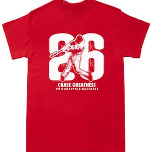 May include: Red t-shirt with a white graphic of a baseball player swinging a bat with the number 26 behind him. The text "CHASE GREATNESS" and "PHILADELPHIA BASEBALL" is below the graphic.
