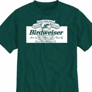 May include: A green t-shirt featuring a white graphic of a beer label that reads "Philadelphia Birdweiser The King of Football Ht on Low Ht on High and watch our Birds Fly World's Greatest Fans Brand Breed Philadelphia Pa"