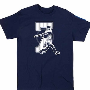 Può includere: Una maglietta blu navy con una grafica bianca di un giocatore di baseball che oscilla una mazza da baseball davanti al numero "7".