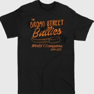 Puede incluir: Camiseta negra con un gráfico naranja de un palo de hockey y un disco. El gráfico dice "The Broad Street Bullies World Champions 1974-1975".