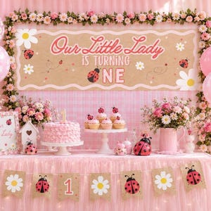 May include: A pink ladybird-themed birthday party setup featuring a banner that reads Our Little Lady is Turning ONE, a ruffled pink cake, cupcakes, floral arrangements in pink pitchers, pink balloons, and a bunting with ladybirds and daisies.
