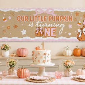 Może przedstawiać: Prostokątny baner z beżowym środkiem i różowymi paskami na brzegach, zawierający napis "OUR LITTLE PUMPKIN is turning ONE" w kolorze pomarańczowym i różowym. Wzór zawiera ilustracje dyń z różowymi kokardkami, jesienne liście i cukierki.
