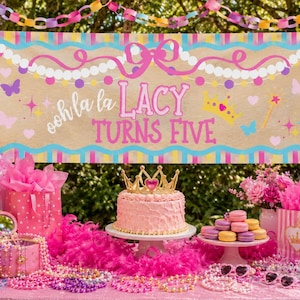 May include: A rectangular banner with the text ooh la la LACY TURNS FIVE, featuring a tan background, pink and blue borders, and illustrations of pearls, butterflies, a crown, and a wand, displayed above a pink cake with a gold crown topper.
