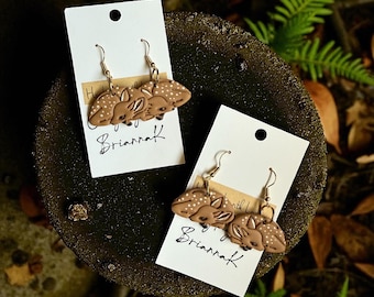 May include: Two pairs of brown deer earrings with white spots and gold-colored hooks are displayed on white cards. The cards have the handwritten name "BriannaK" and are set against a dark, textured background with greenery.