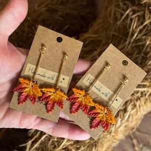 May include: A pair of gold-toned dangle earrings featuring autumn-colored maple leaf charms. Each earring has a small gold ball at the top and a delicate chain suspending the leaf. The text "Handmade with Love" is printed on the earring card.