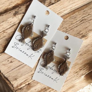 May include: A pair of brown fish-shaped earrings with silver hooks. The earrings are on a white card with the text "Clayday with Brianrak" written on it.
