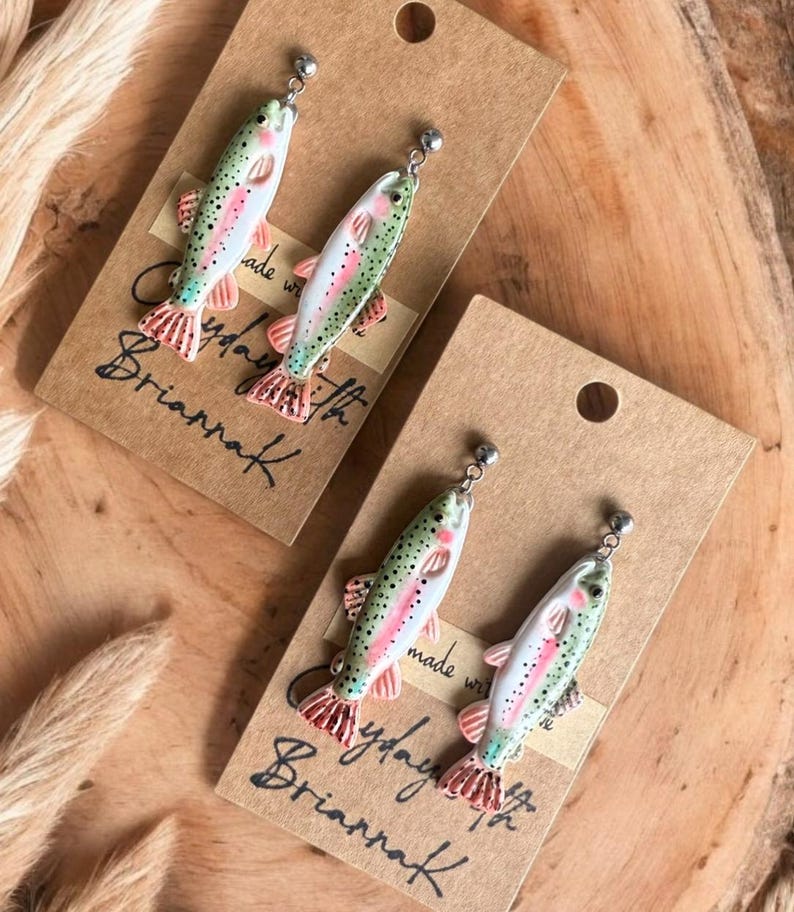 May include: A pair of silver stud earrings featuring a hand-painted trout design. The trout are green, pink and white with black spots. The earrings are displayed on a brown card with the text 'Made with love by Brianna' written on it.