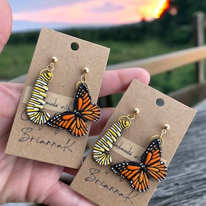 K&ouml;nnte beinhalten: Ein Paar handgefertigte Ohrringe mit einem Monarchfalter-Motiv. Die Ohrringe sind aus Polymer Clay gefertigt und haben goldfarbene Haken. Die Ohrringe werden auf einer braunen Karte mit dem Text "Handmade with Love by Briarraf" pr&auml;sentiert.
