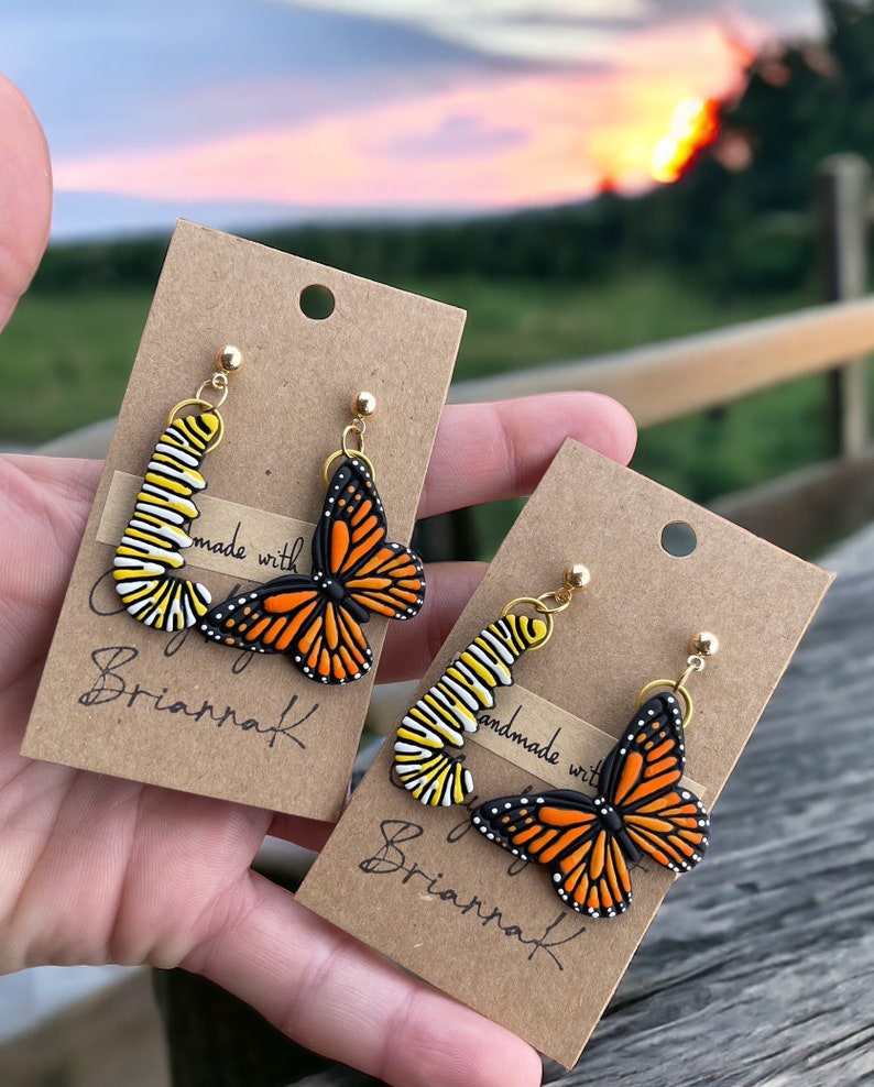 K&ouml;nnte beinhalten: Ein Paar handgefertigte Ohrringe mit einem Monarchfalter-Motiv. Die Ohrringe sind aus Polymer Clay gefertigt und haben goldfarbene Haken. Die Ohrringe werden auf einer braunen Karte mit dem Text "Handmade with Love by Briarraf" pr&auml;sentiert.
