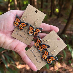 May include: Two pairs of gold butterfly earrings with black and orange wings. The earrings are on brown cards with the text "Butterfly Briarshaft" written on them.
