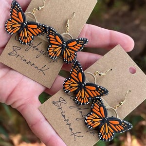 May include: A pair of gold butterfly earrings with black and orange wings. The earrings are displayed on a brown card with the text "Play with BriannaK" written on it.